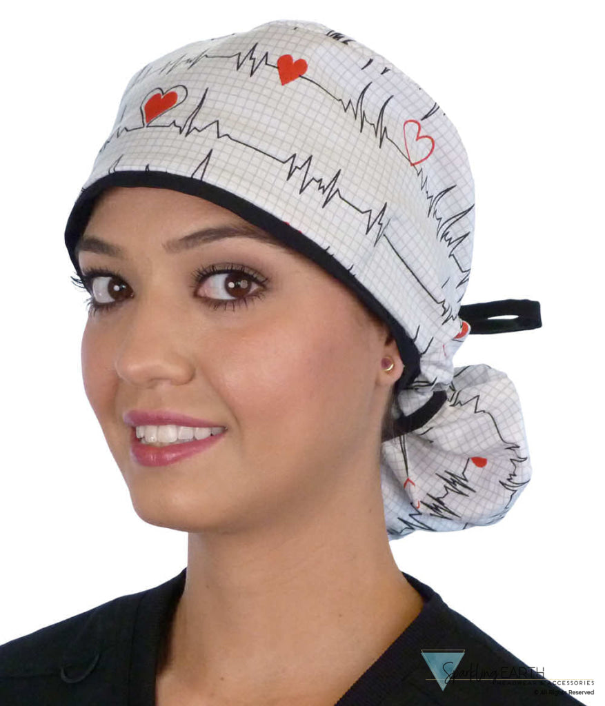 Front facing image of a woman in scrubs wearing a Big Hair Ponytail Scrub Cap with a Heartbeats on White pattern, and black ties to hold the scrub cap in place.